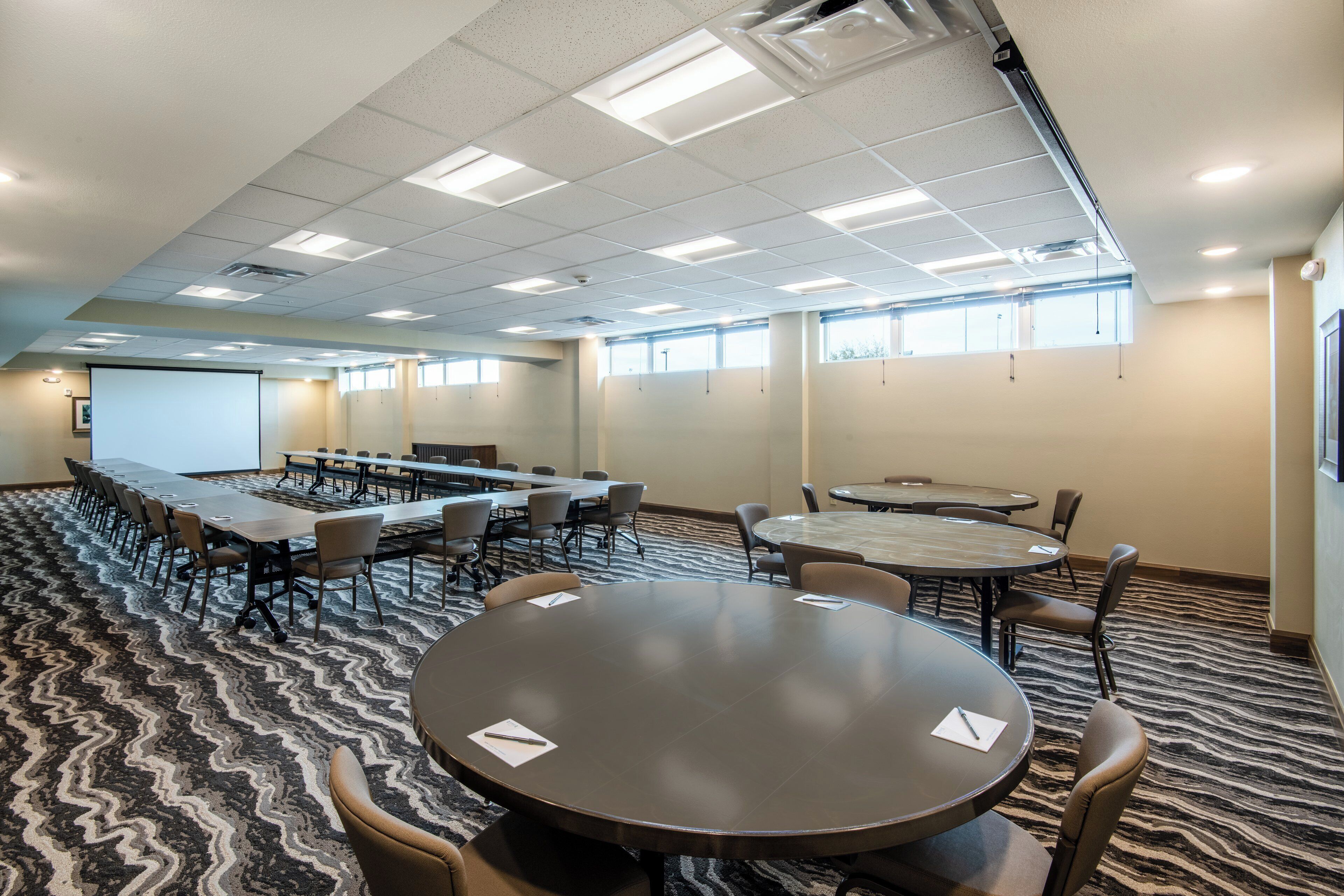 A conference room with a long table and smaller round tables, equipped with chairs and a projector screen.
