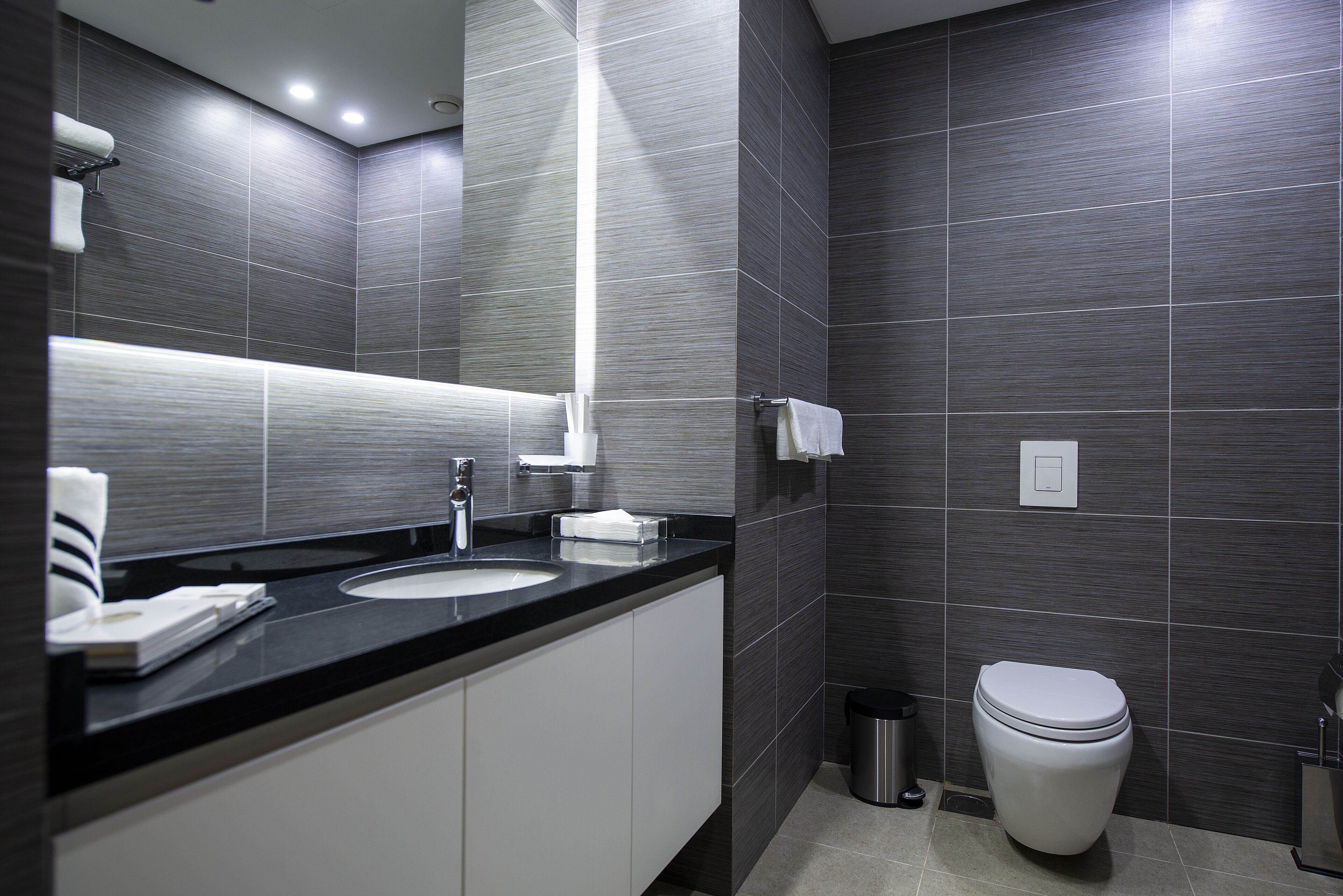 Un baño moderno con paredes de azulejos oscuros, una encimera negra, un lavabo blanco y un inodoro.