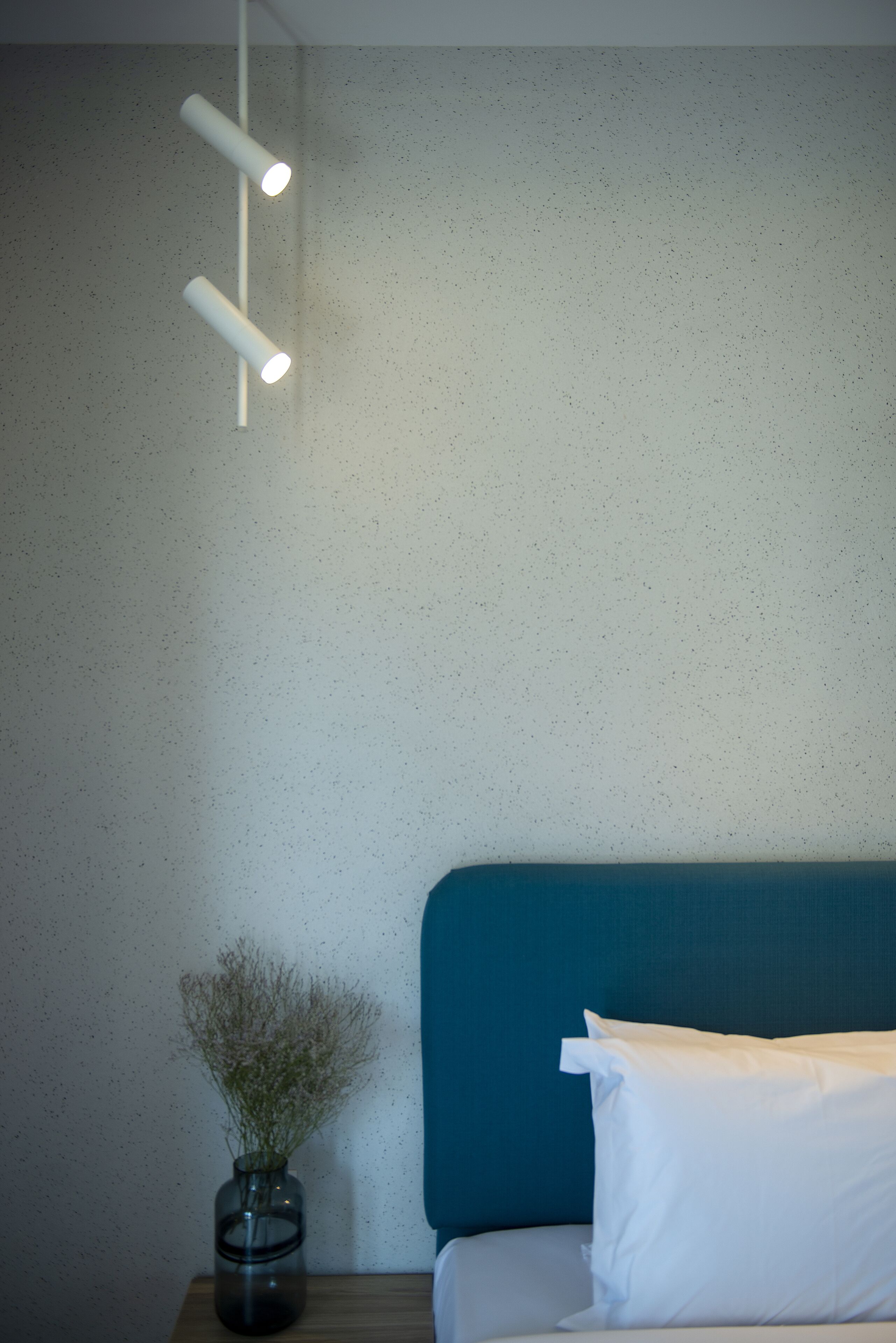 Un dormitorio con un cabecero color turquesa, una almohada blanca y un jarrón con flores secas sobre una mesita de noche de madera.