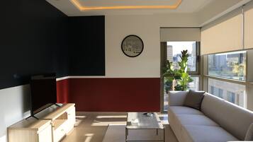 Una sala moderna con sofá, televisor y vistas a la ciudad a través de amplias ventanas.