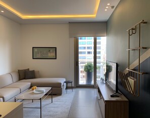 A modern living room with a sofa, coffee table, and a TV on a wooden stand. There is a framed picture on the wall and a balcony with potted plants. - ZUR Studios & Suites (Beirut)