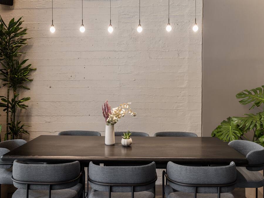 Un comedor con una mesa rectangular, seis sillas grises, focos colgantes y plantas en macetas.
