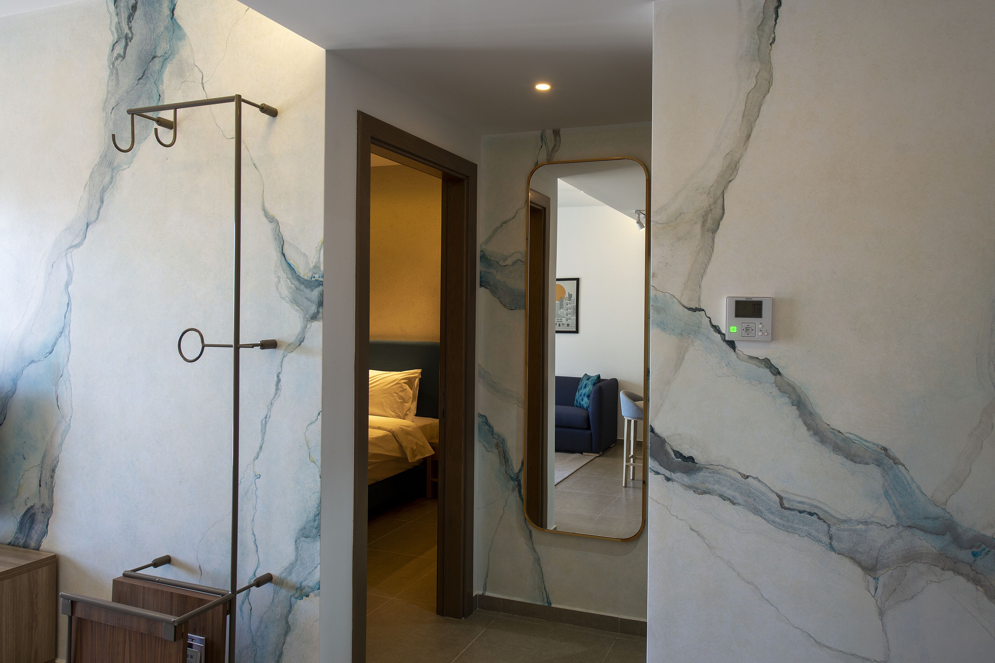A hotel room with a marble wall, a bed, a sofa, and a bathroom with a mirror and a towel rack.