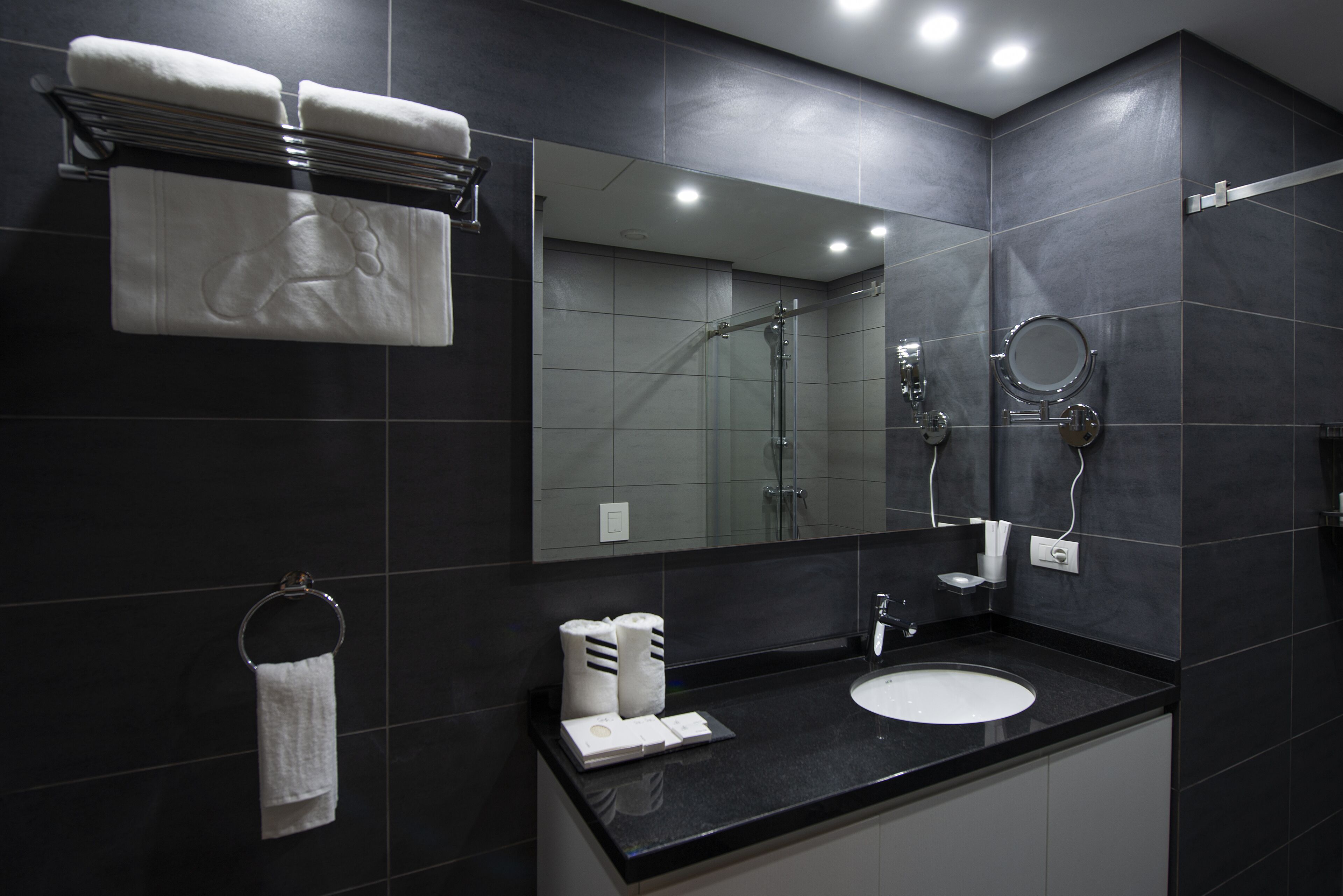 A modern bathroom with dark tiles, a black countertop, a white sink, a glass shower enclosure, and a large mirror.
