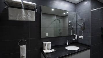 A modern bathroom with dark tiles, a black countertop, a white sink, a glass shower enclosure, and a large mirror.