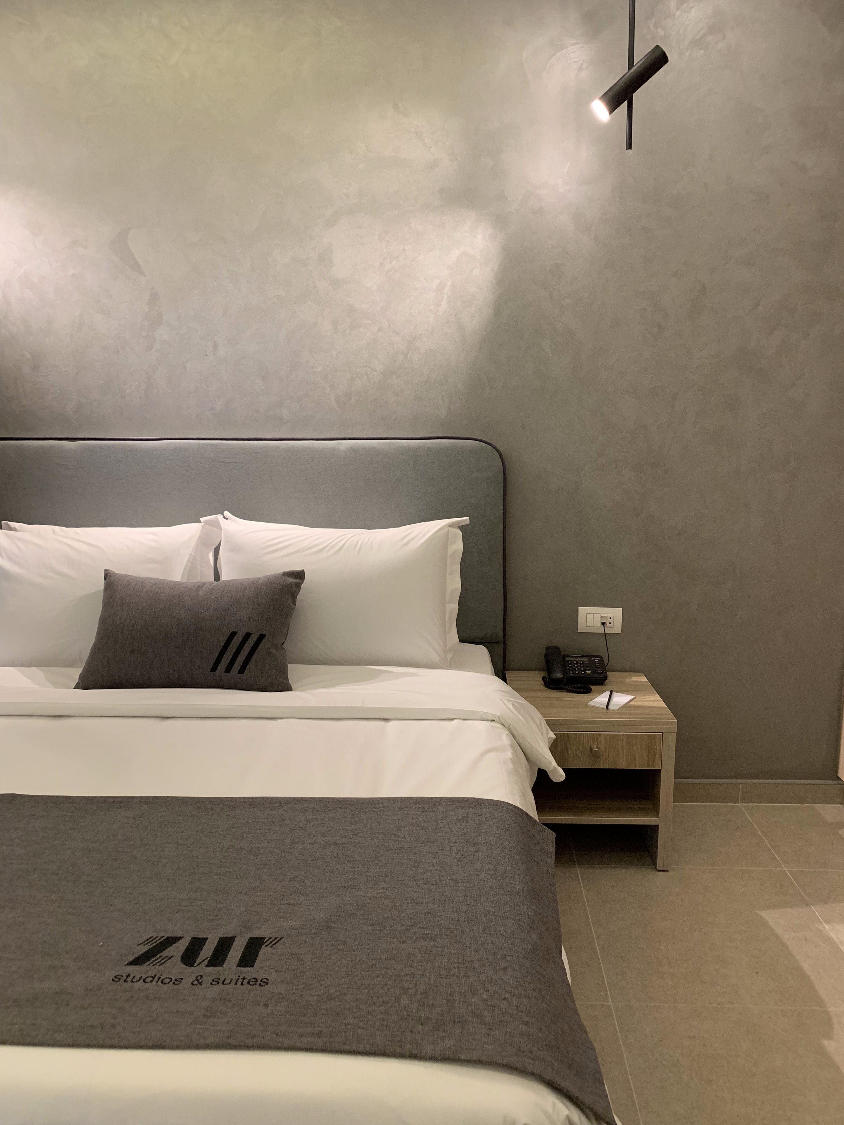 A modern bedroom with a bed, pillows, a bedside table with a phone, and a wall-mounted light.