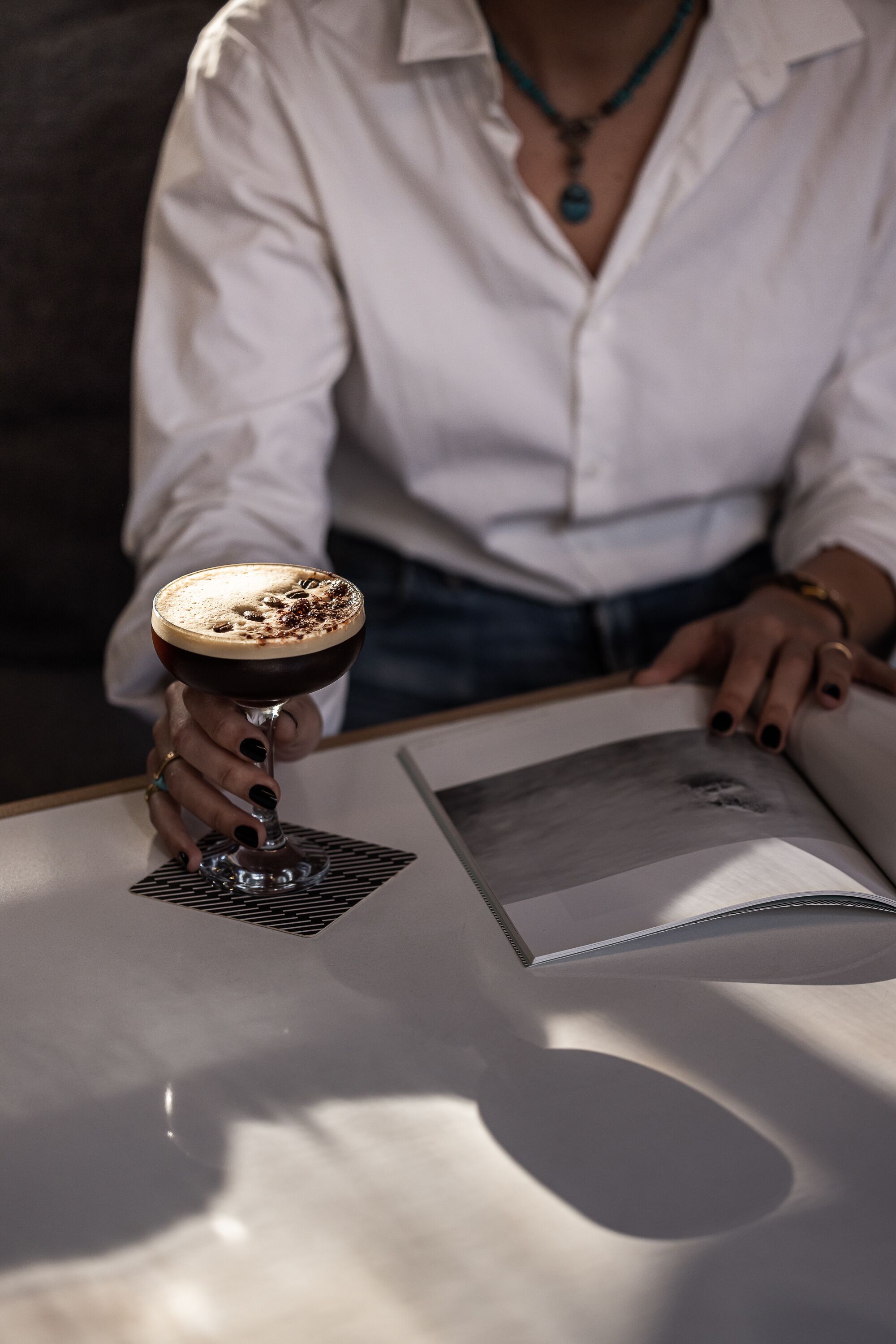 A person holding a cocktail glass on a table with a magazine.