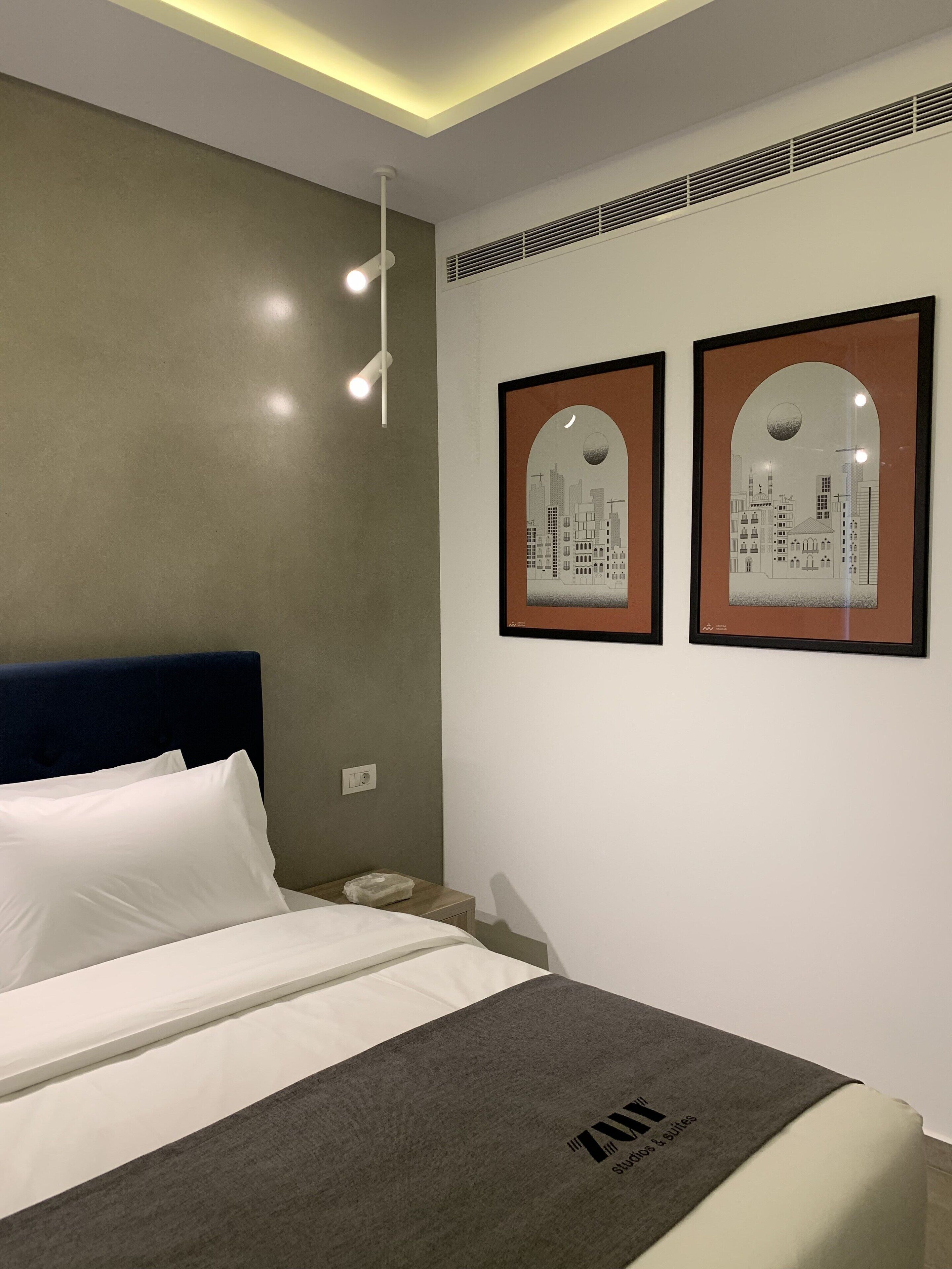 Een hotelkamer met een bed, een nachtkastje, twee ingelijste foto’s aan de muur en een plafondlamp.