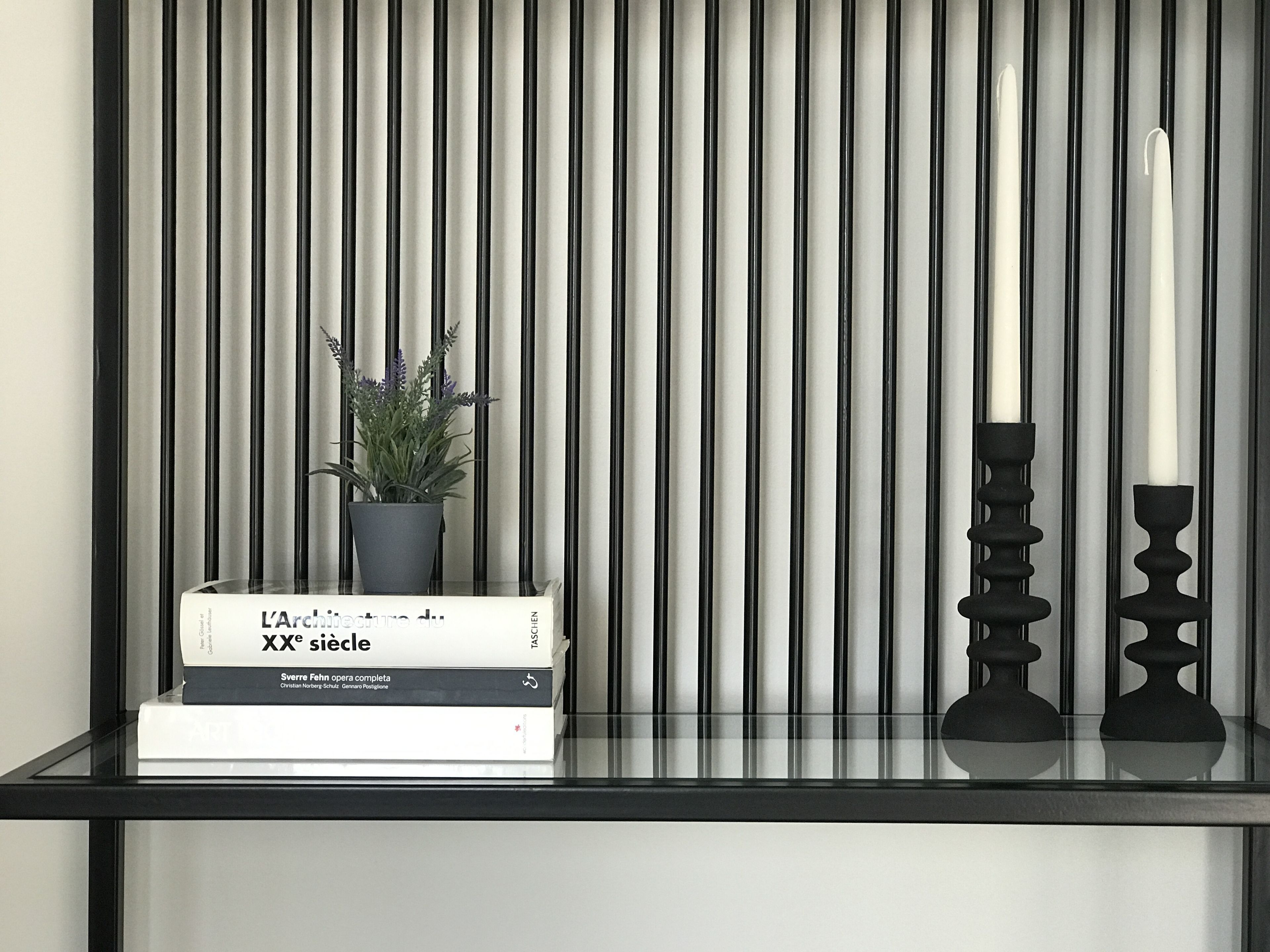 A modern, minimalist shelf with a potted plant, a stack of books, and two candle holders against a striped background.