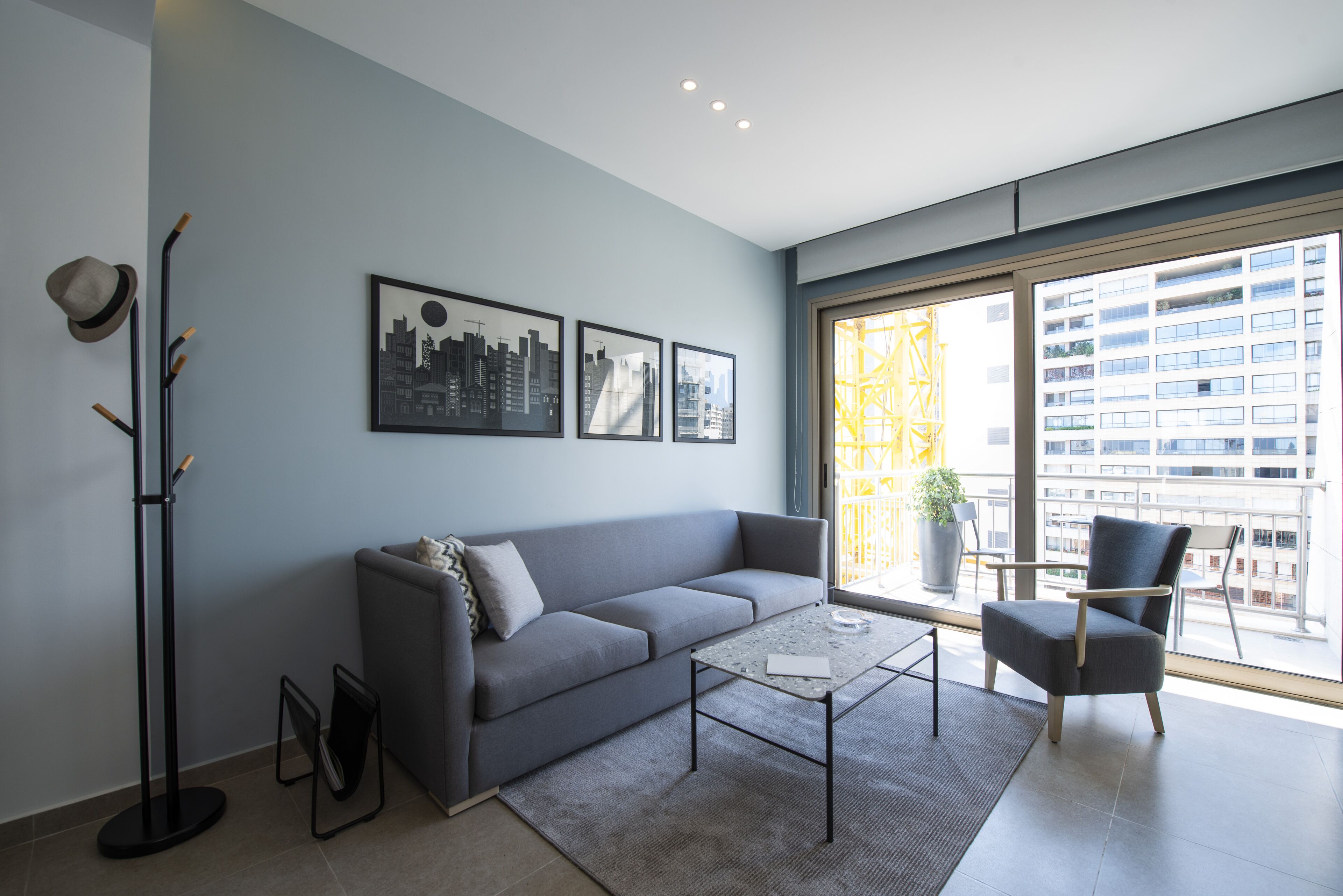 Un salon moderne avec un canapé gris, une table basse et un fauteuil. Trois tableaux encadrés ornent le mur, ainsi qu’une lampe sur pied et une grande fenêtre donnant sur des immeubles.