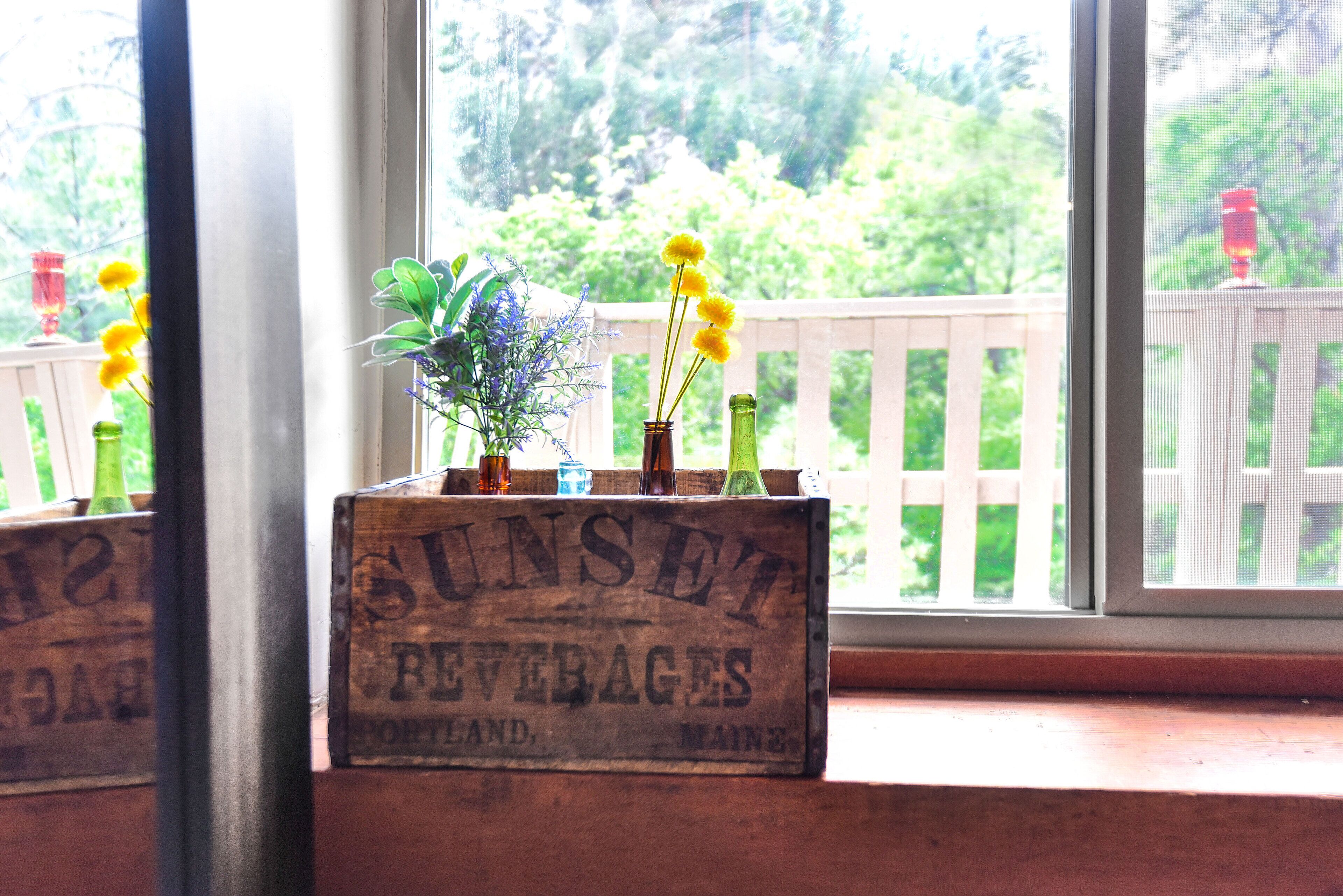 A wooden crate labeled "SUNSET BEVERAGES" with a potted plant and vases on a windowsill.