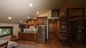 Una cocina compacta con armarios de madera, un refrigerador de acero inoxidable y un microondas. Hay una zona de comedor con una mesa y sillas, y una escalera que lleva a una zona superior.