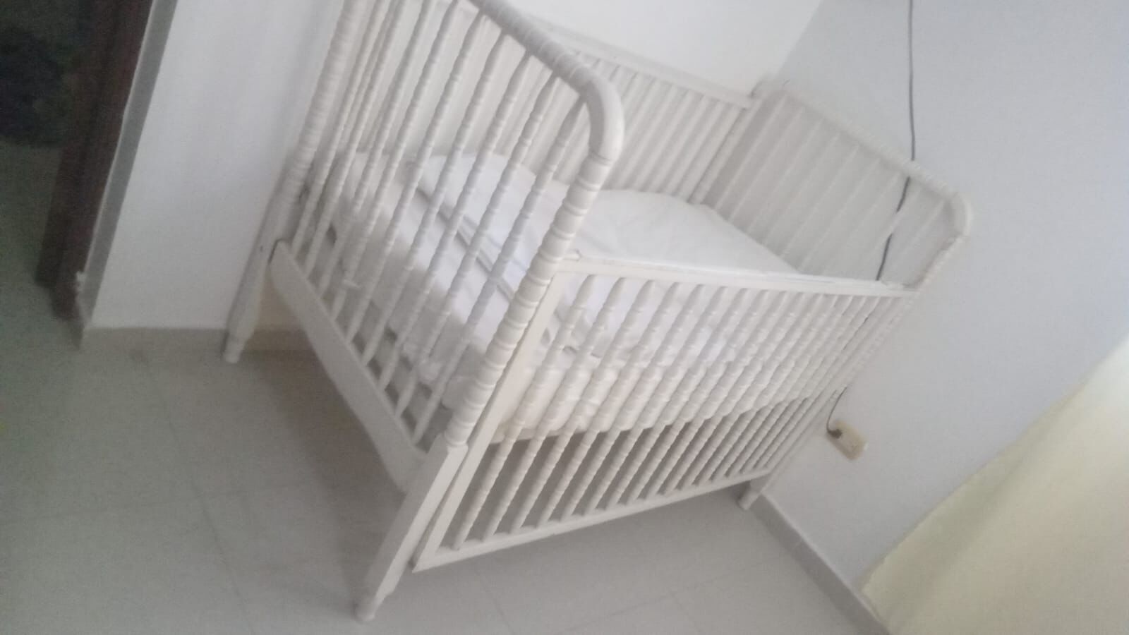 Een wit wiegje met een gebogen hoofdeinde, geplaatst tegen een witte muur in een kamer met een betegelde vloer.