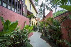 Uma trilha estreita ladeada por plantas tropicais exuberantes que leva a um edifício com fachada vermelha e uma varanda.