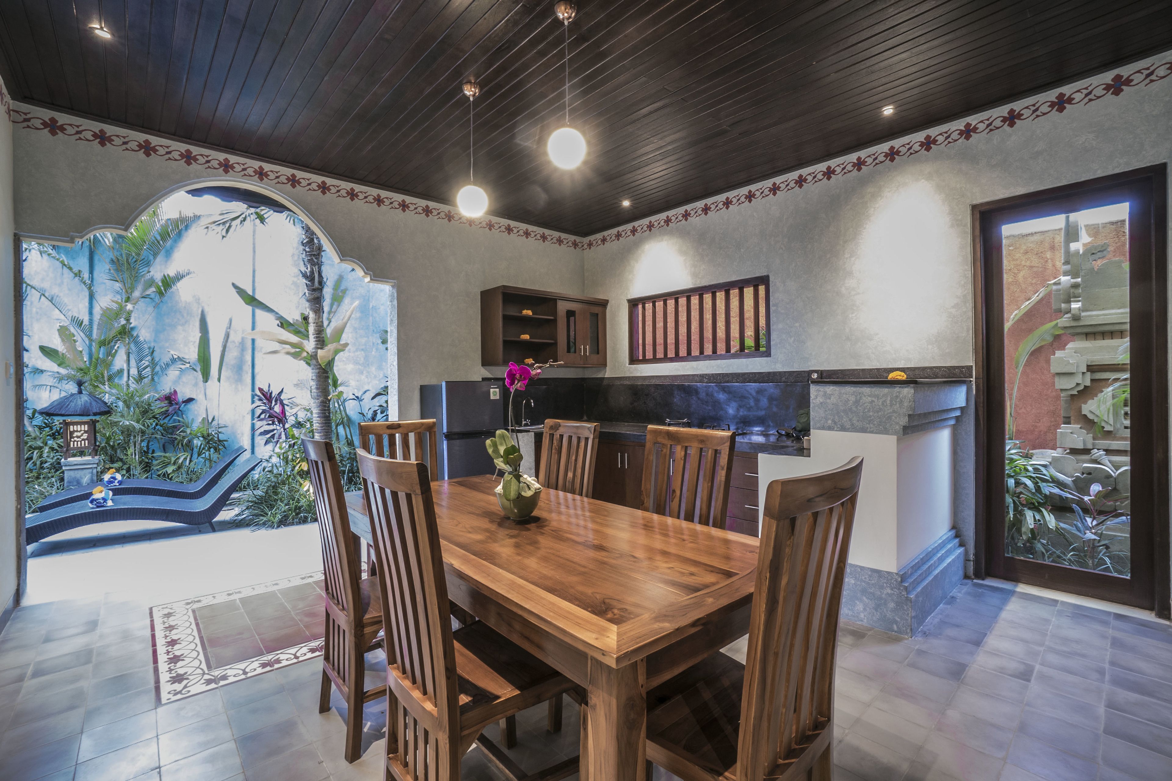 A spacious dining area with a wooden table and chairs, a kitchen with modern appliances, and a view of a tropical garden through an arched doorway.