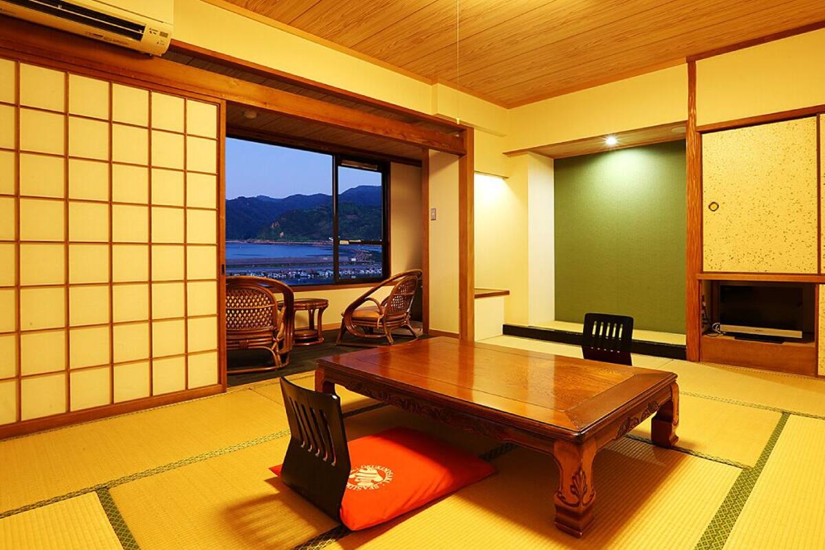 Ein traditioneller japanischer Raum mit Tatami-Böden, einem Holztisch und Stühlen. Eine Schiebetür führt zu einem Balkon mit Blick auf das Meer und die Berge.