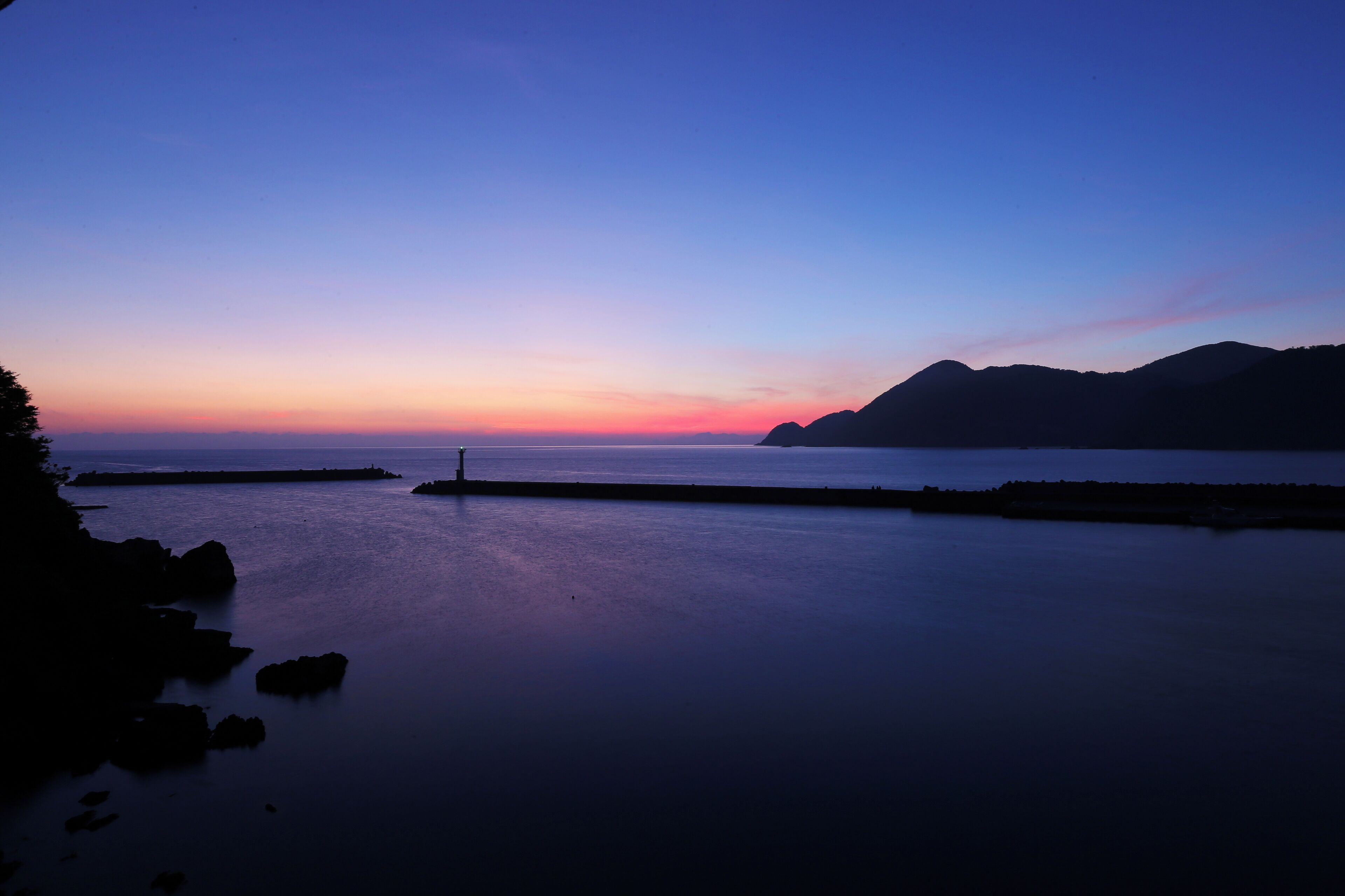 Un paisaje costero con mar tranquilo, un muelle y montañas distantes al atardecer.