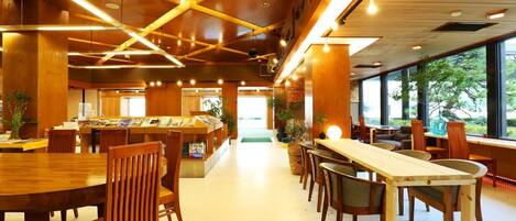 A spacious indoor area with wooden tables and chairs, a reception desk, and large windows offering a view of greenery outside.