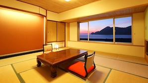A traditional Japanese room with tatami flooring, a low wooden table, and a view of a lake and mountains at sunset. - Okukinosaki Seaside Hotel (Toyooka)