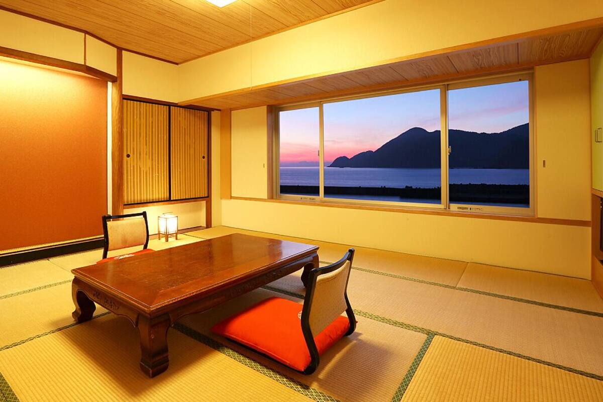 Ein traditioneller japanischer Raum mit Tatami-Böden, einem niedrigen Holztisch und Blick auf einen See und Berge bei Sonnenuntergang.
