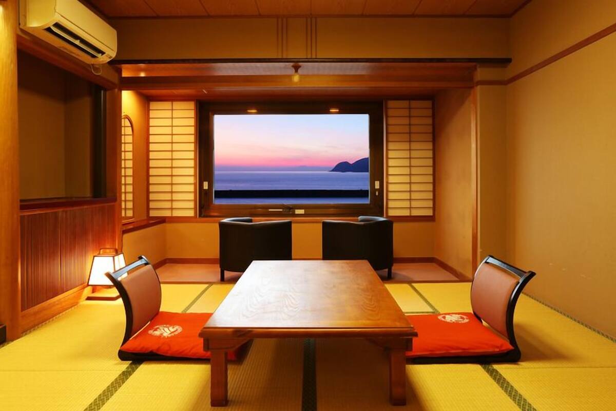Ein traditioneller japanischer Raum mit Tatami-Boden, einem niedrigen Tisch und einem Fernsehbildschirm, das einen Sonnenuntergang zeigt.