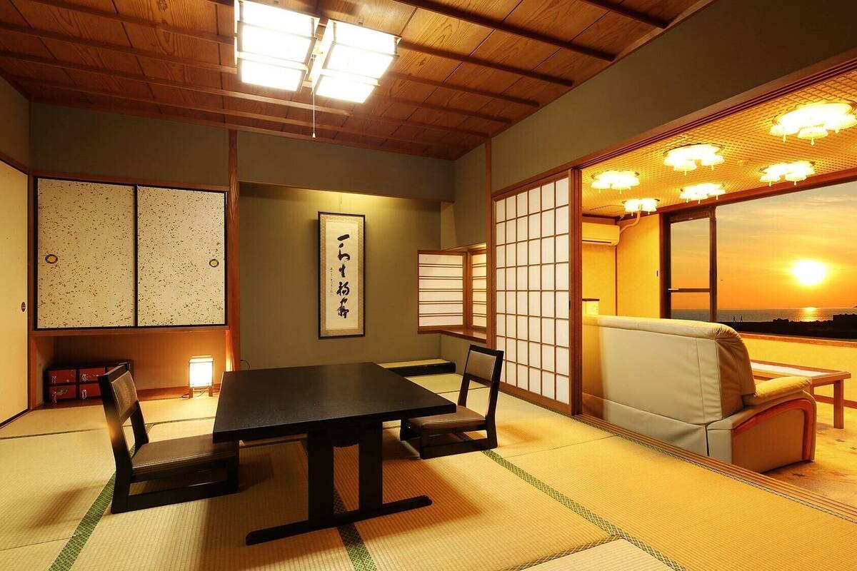 Ein traditioneller japanischer Raum mit Tatami-Boden, einem niedrigen Tisch, einer gepolsterten Sitzbank, Schiebetüren und Blick auf den Sonnenuntergang.