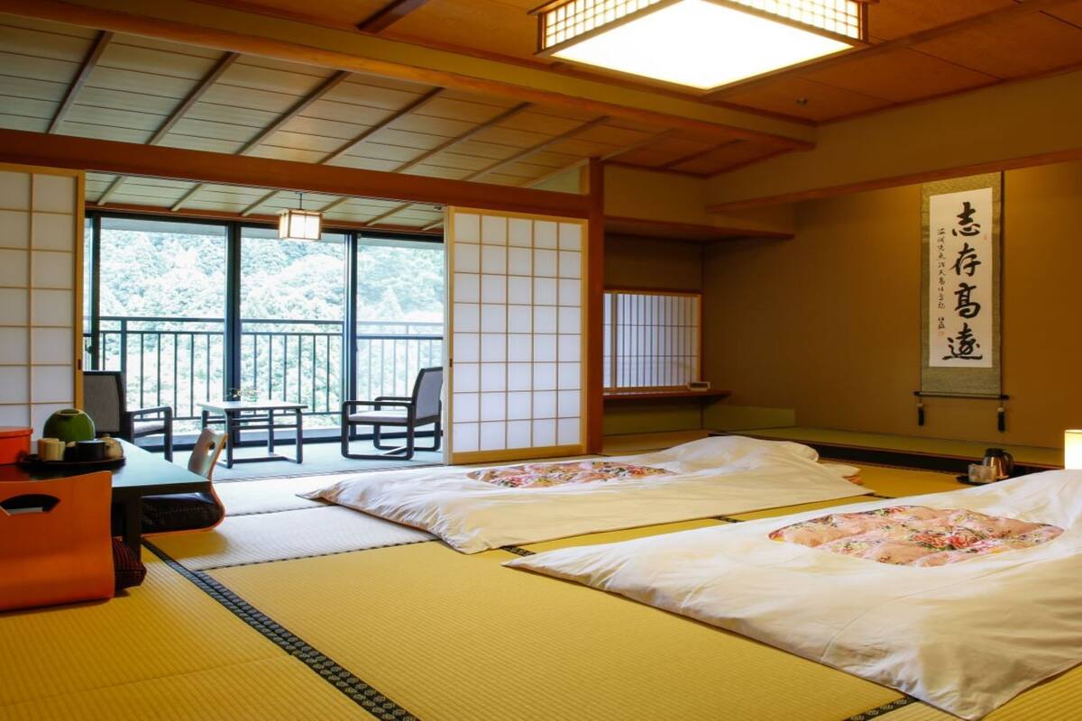 A traditional Japanese room with tatami mats, two beds with floral bedding, a sliding door, a table with a teapot, and a view of greenery outside.
