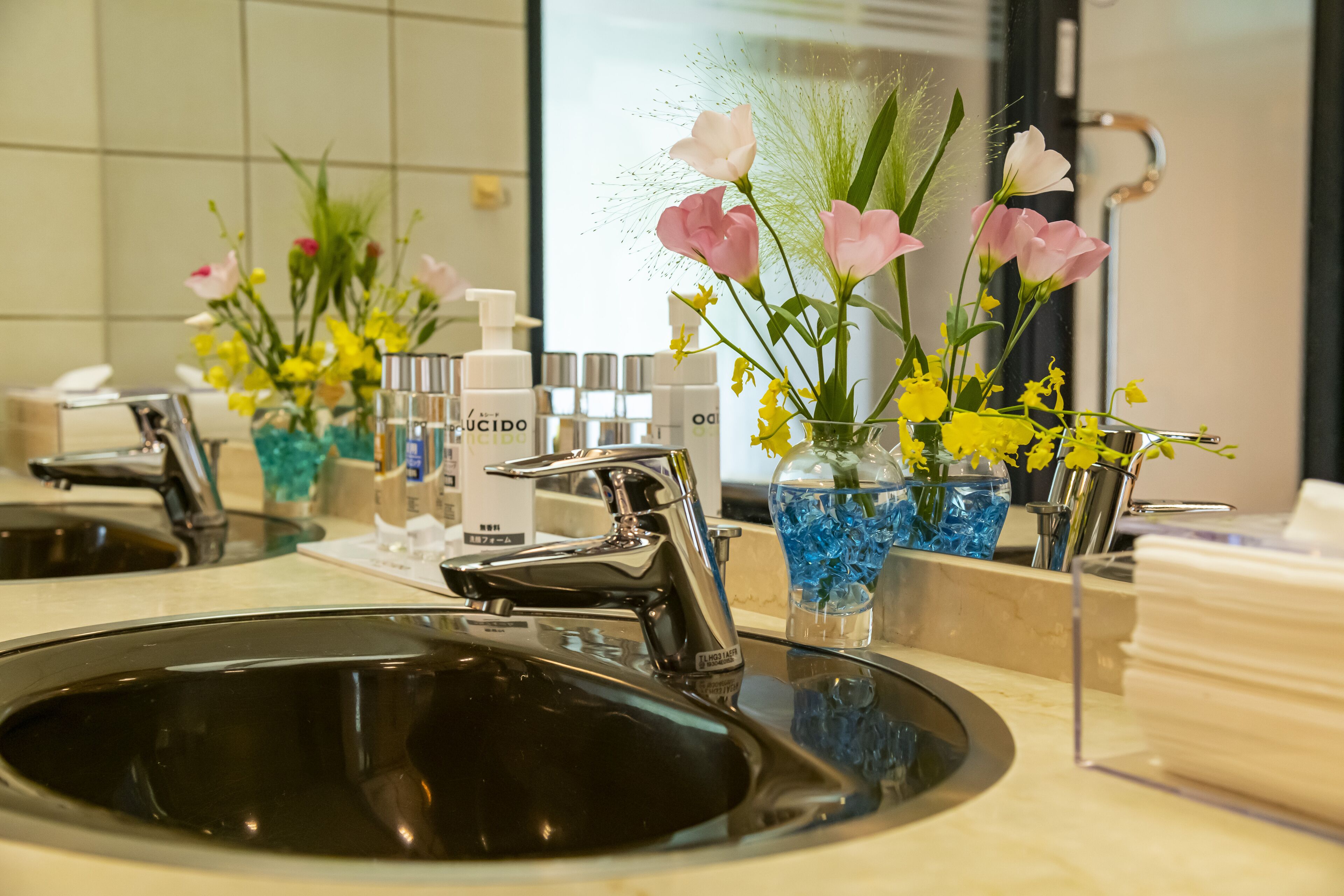 Un lavamanos de baño con una llave moderna, un espejo y dos jarrones con flores.