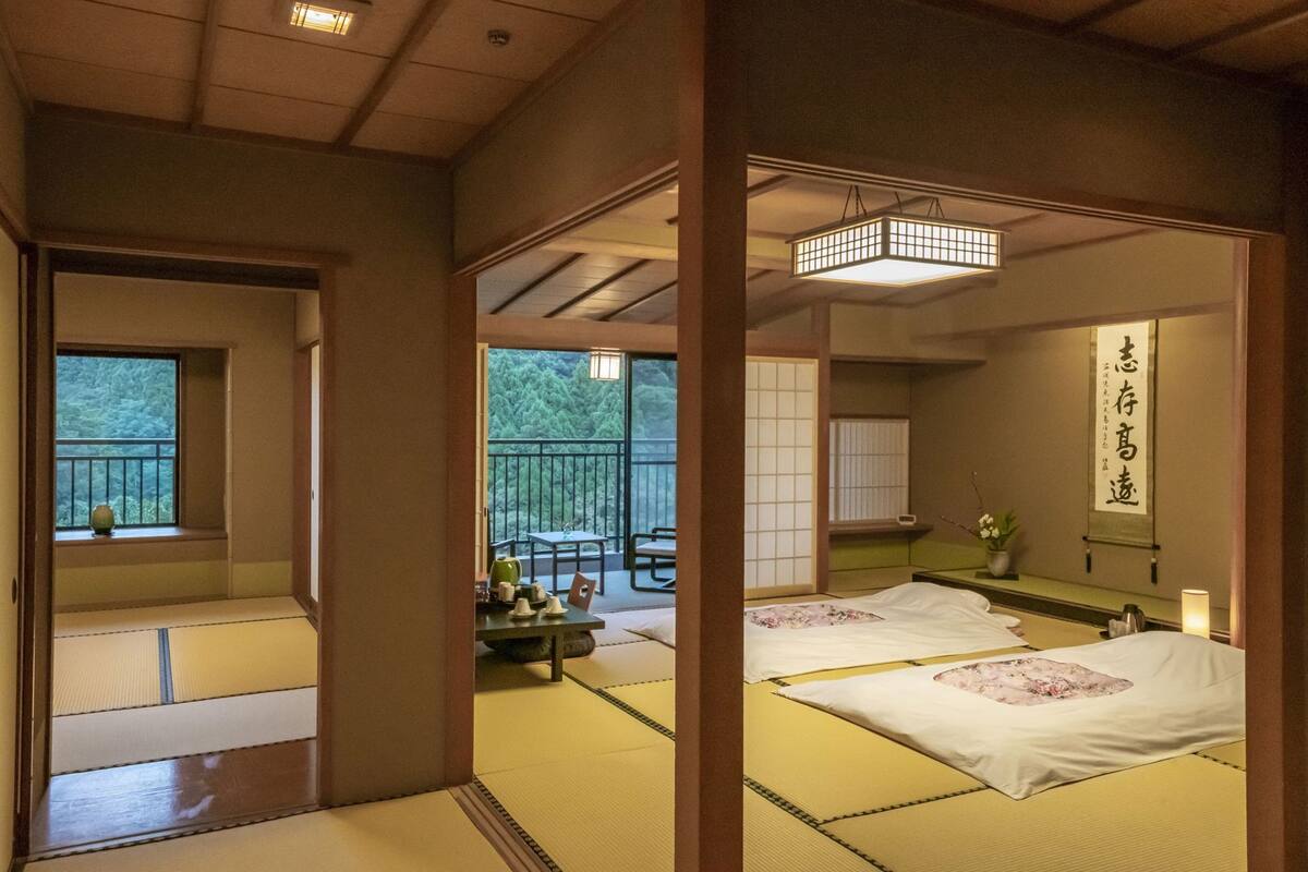 A traditional Japanese room with tatami flooring, a sliding door, a balcony with a view of trees, and a hanging lantern.