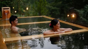 A couple relaxing in a clear, rectangular pool with wooden edges, surrounded by lush greenery and ambient lighting.