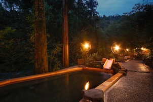 Un espace de sources chaudes doté d’une terrasse en bois, de bancs en pierre et d’une piscine entourée d’arbres.