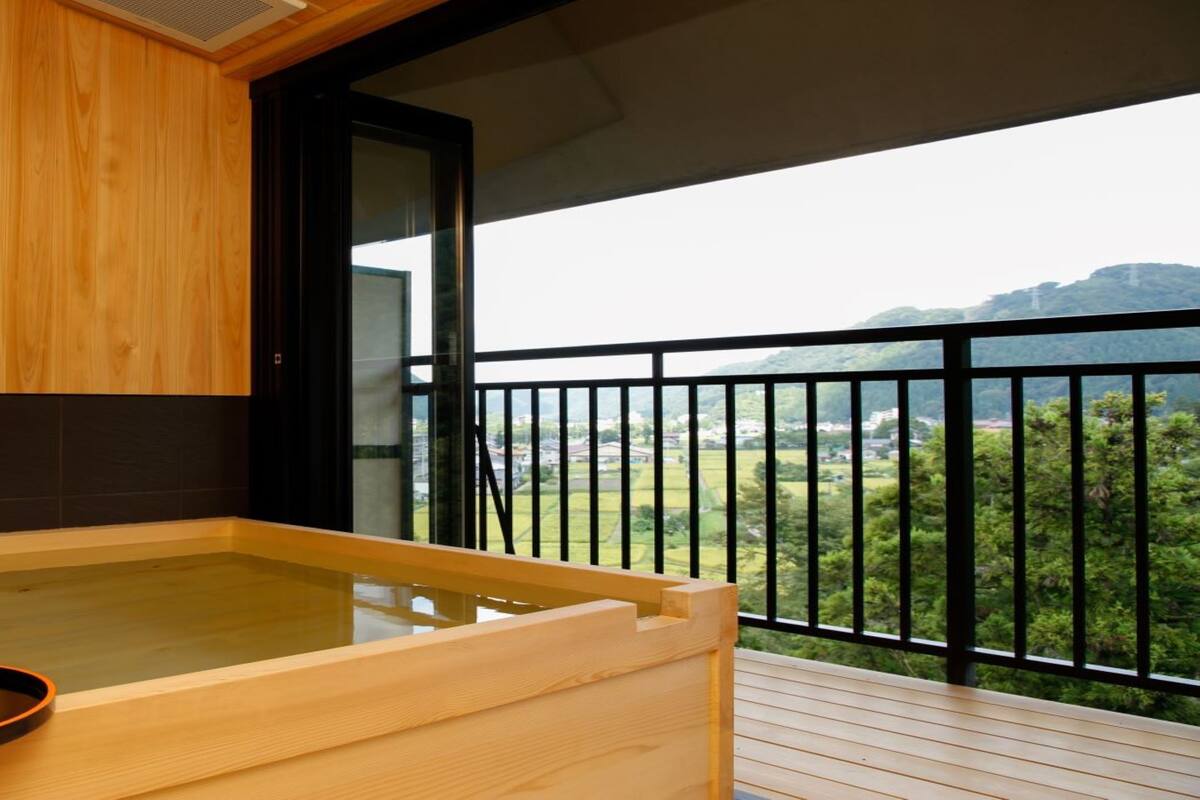A wooden spa tub with a tray and a small vase on a balcony with a view of greenery and hills.