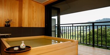 Tinaja de madera tipo spa con una bandeja y un jarrón pequeño en un balcón con vistas a follaje y colinas.