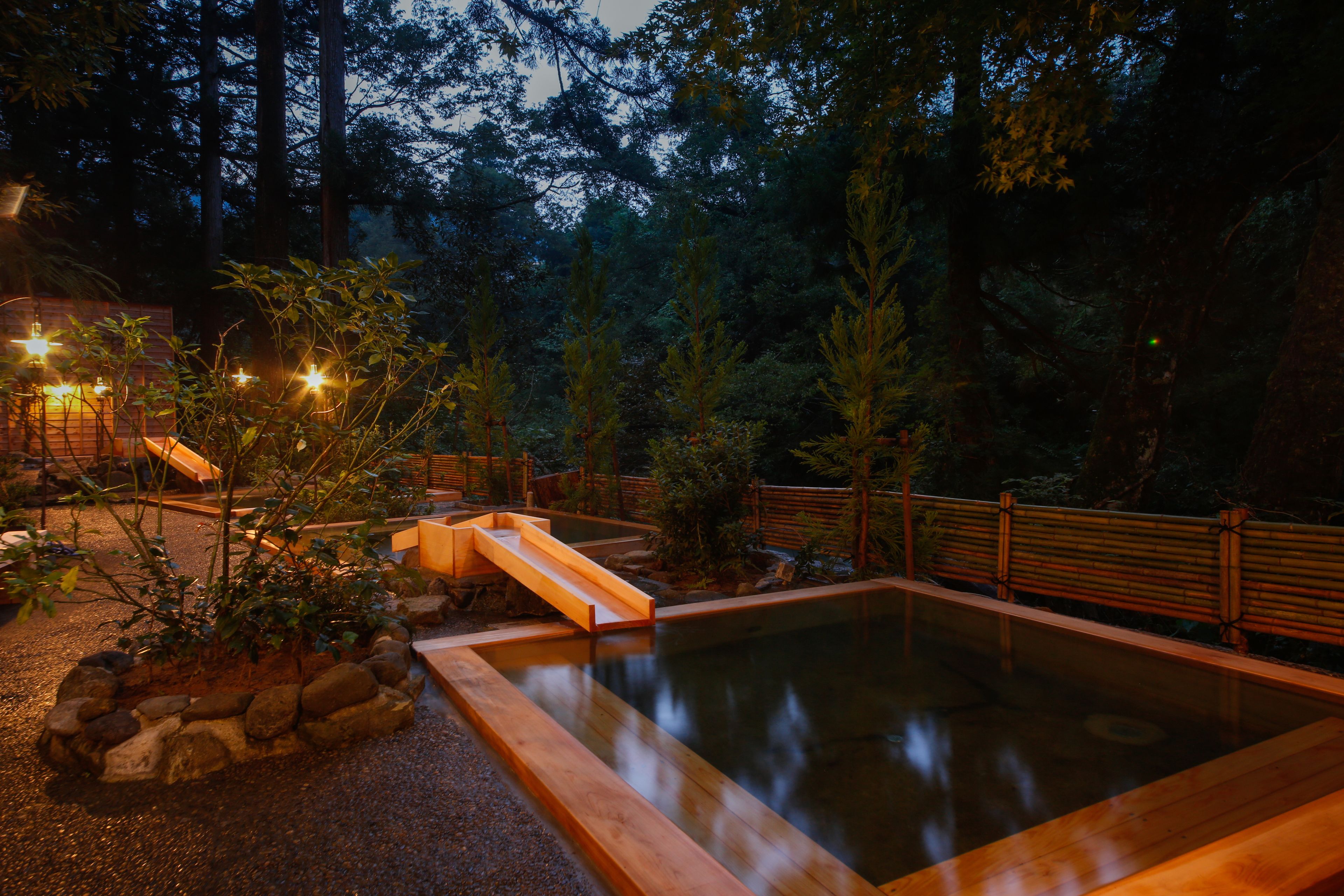 A serene outdoor pool area with a wooden deck, surrounded by rocks and plants, and a wooden bridge leading to the pool.