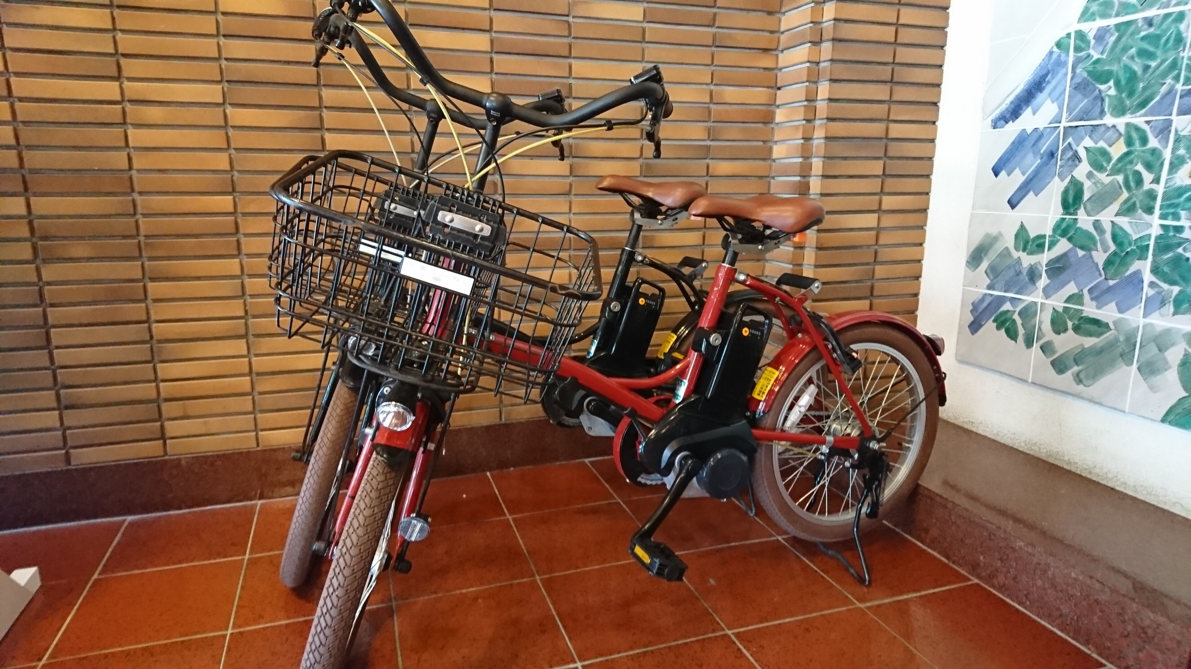 A red bicycle with a basket, parked indoors with a tiled floor and a brick wall in the background.