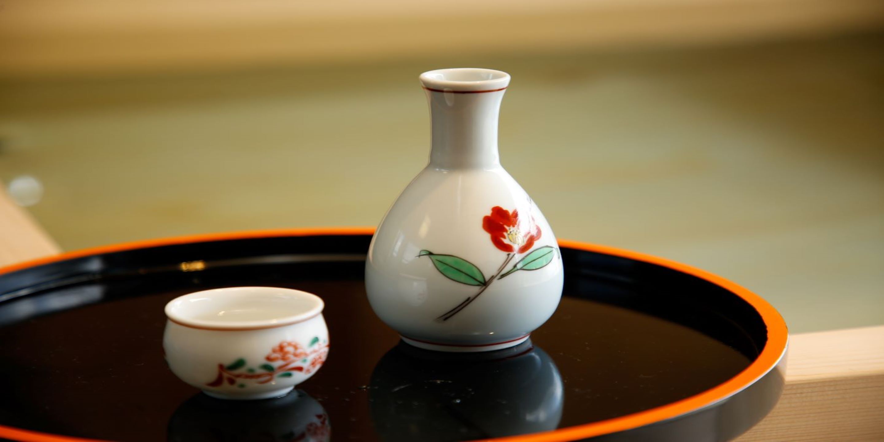 A white ceramic vase with a floral design and a smaller bowl on a black tray with an orange rim.