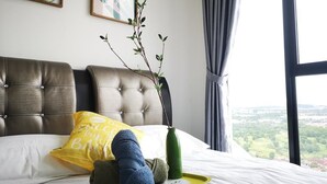 A bedroom with a tufted headboard, a bed with white bedding, a yellow pillow, a green vase with a branch, and a window with a view of greenery. - YOYO Homestay (Johor Bahru)