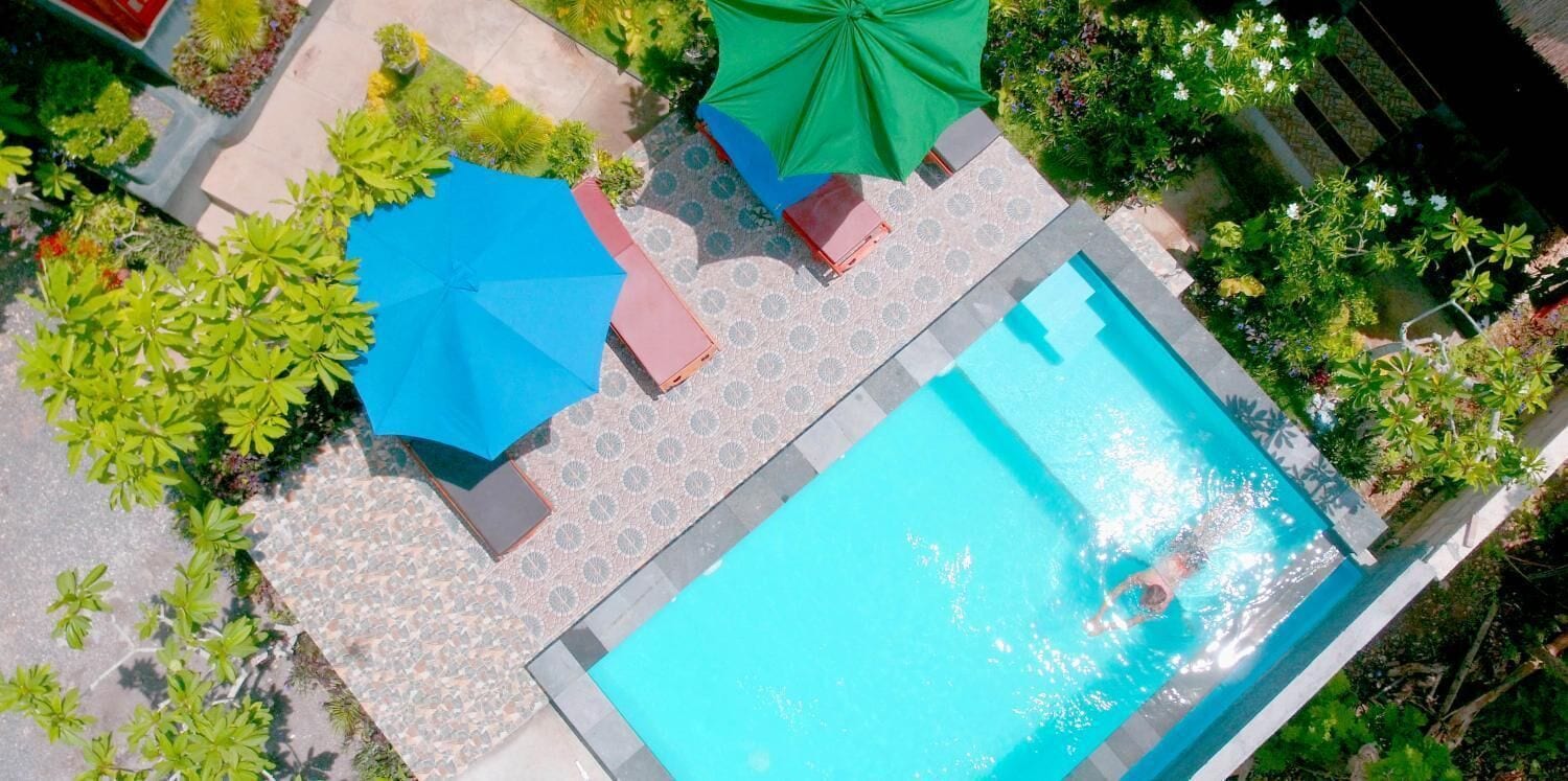 Una piscina con borde de baldosas, rodeada de vegetación y dos sombrillas azules.