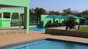 A swimming pool with a green fence and umbrellas, surrounded by a landscaped area with a small tree and outdoor seating. - Mashamba Country House (Giyani)