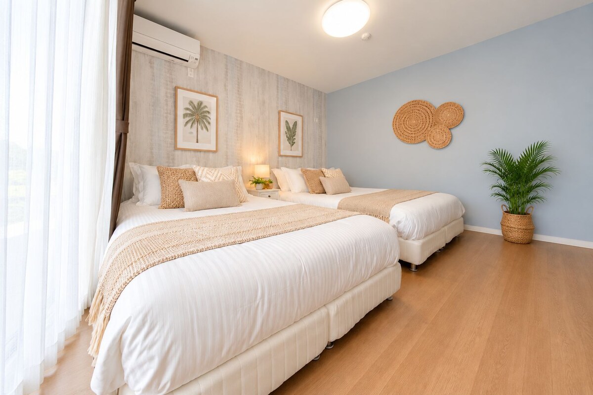 A hotel room with two beds, a large window with sheer curtains, a wall-mounted air conditioner, and a woven wall decoration.