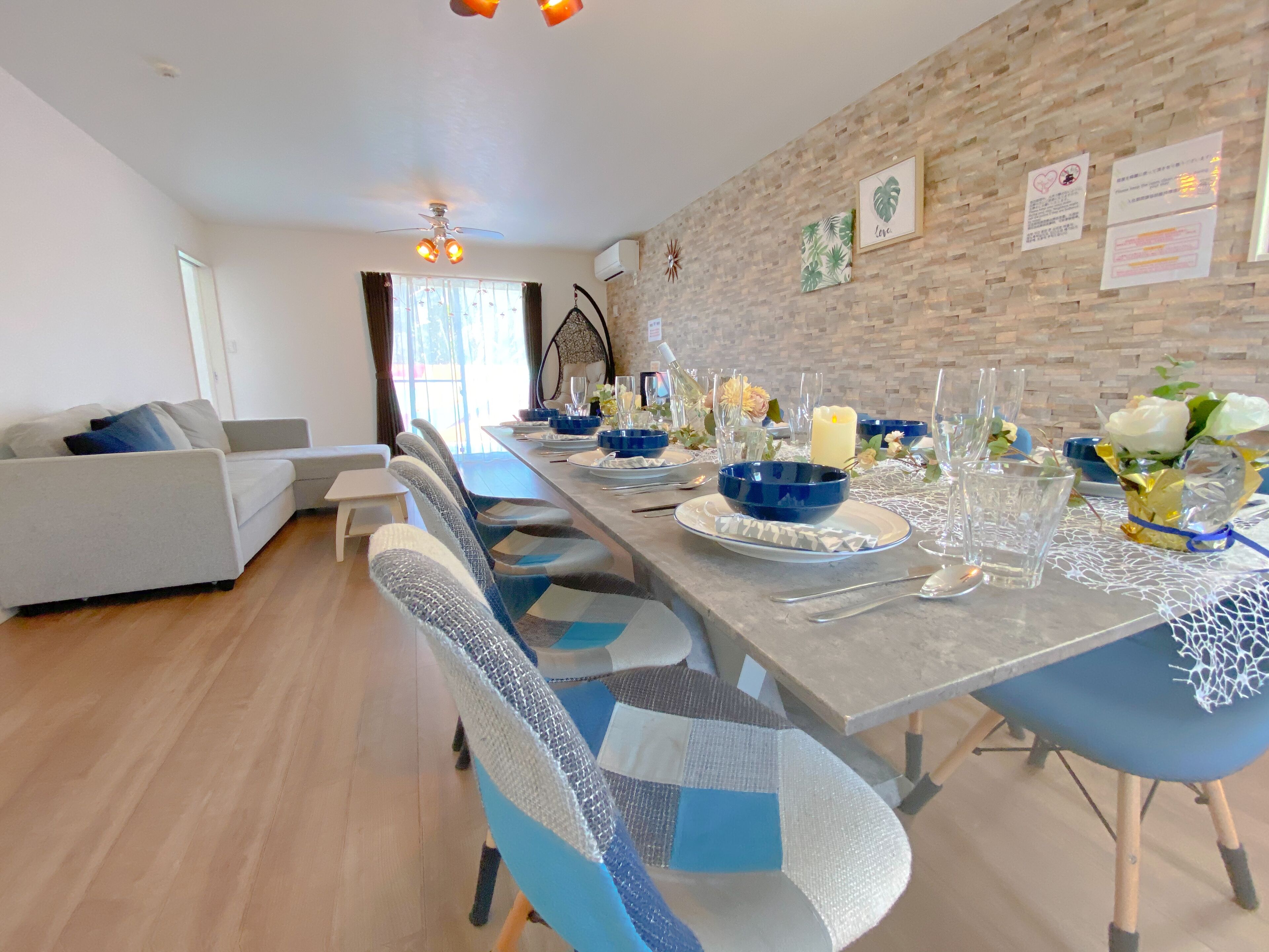 Een moderne eetruimte met een lange tafel gedekt voor zes personen, met blauwe en witte stoelen, helder glaswerk en decoratieve elementen zoals kaarsen en bloemen.
