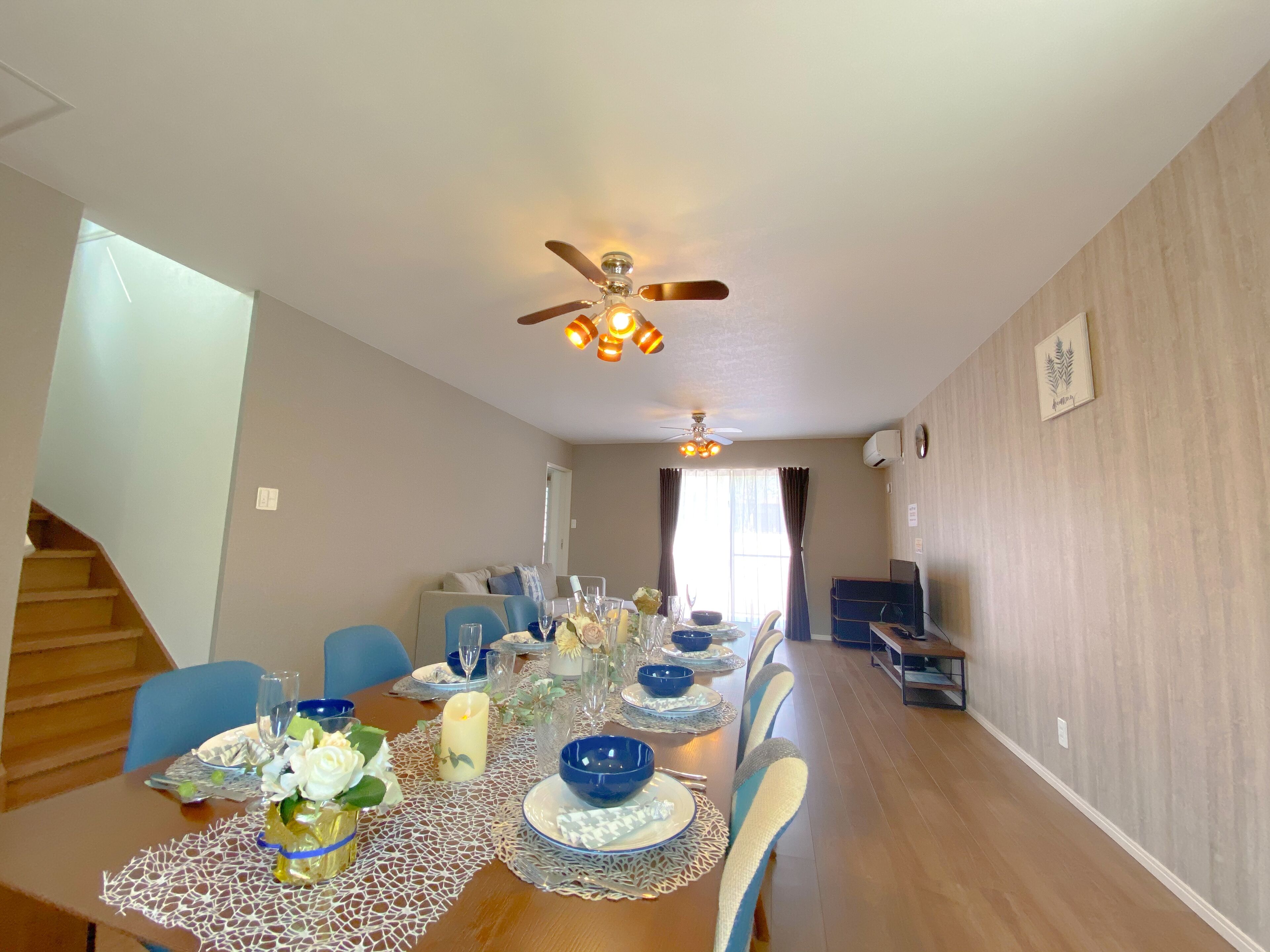 A dining area with a long table set for six, a ceiling fan, and a staircase leading to another room.