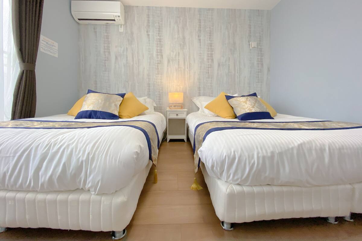 Une chambre d’hôtel avec deux lits, chacun agrémenté de linge de lit blanc et de touches de bleu et d’or. On y trouve un climatiseur mural, une table de chevet avec une lampe et un plancher en bois.