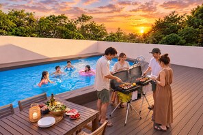 A poolside barbecue with people grilling and swimming, surrounded by a wooden deck and a sunset view. - Grandioso Okinawa Pool Villa Onna 4 (Onna)
