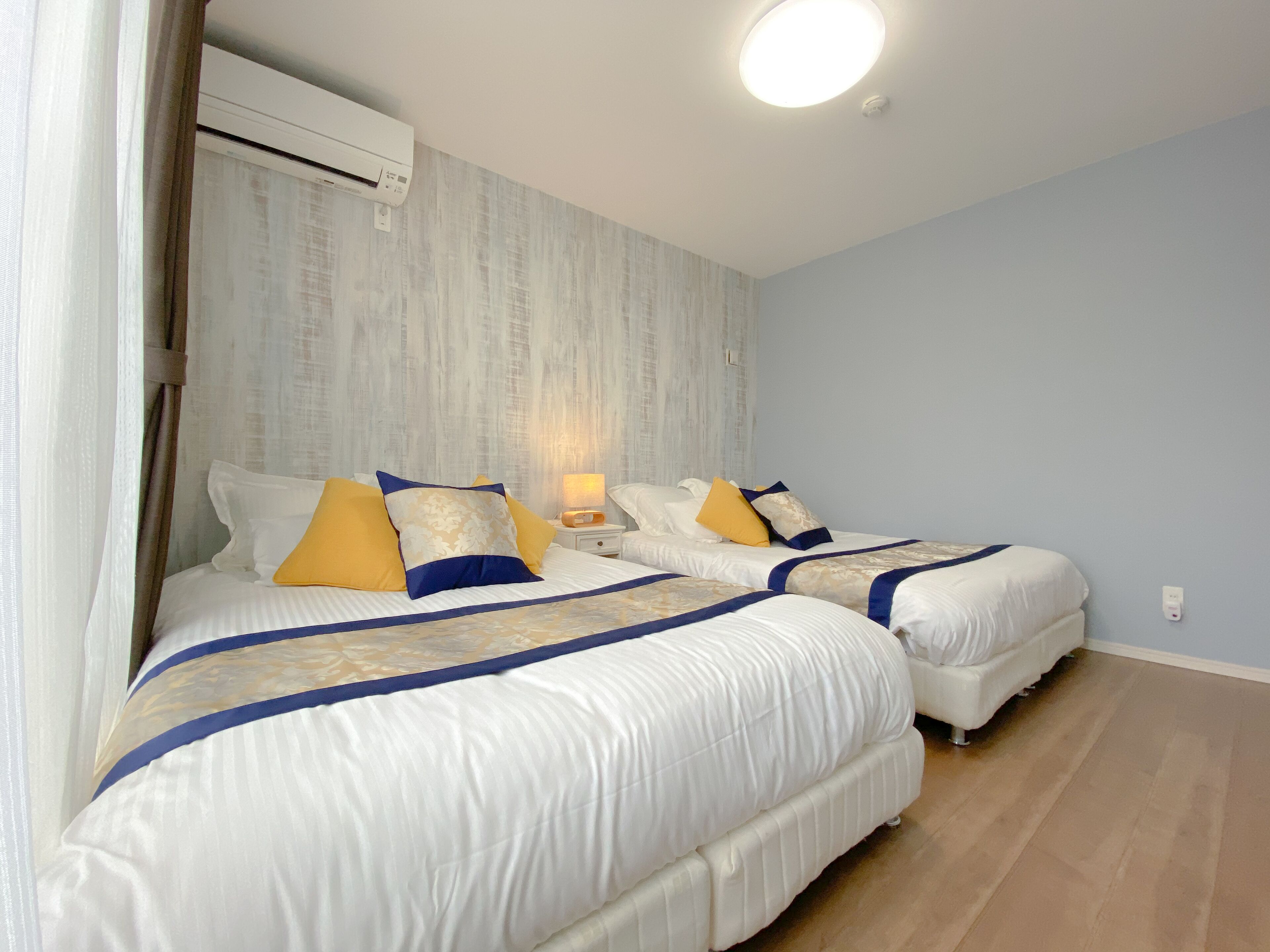 A hotel room with two beds, a ceiling fan, an air conditioning unit, and a bedside lamp.