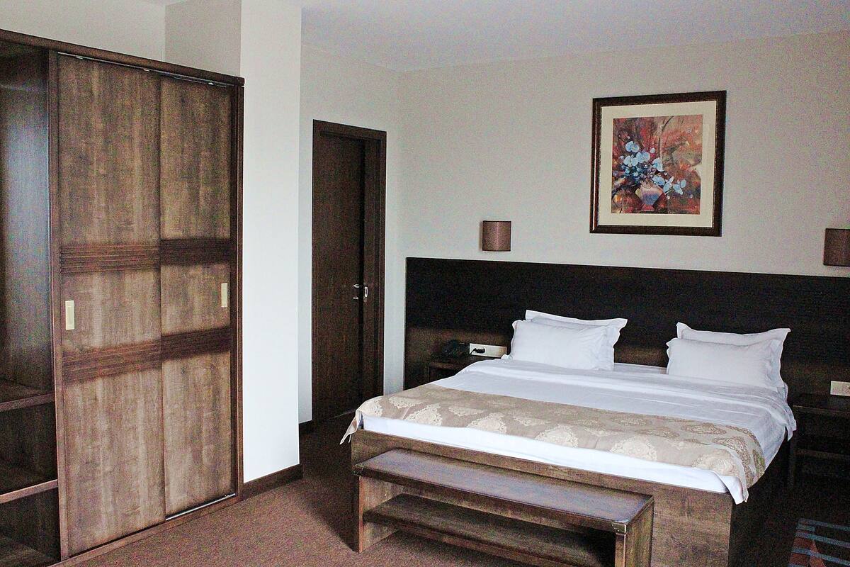 Habitación de hotel con una cama grande, un armario de madera, un banco, una mesita de noche y un cuadro en la pared.