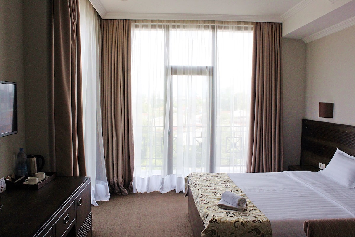 Habitación de hotel con una cama grande, un escritorio con televisor y vistas al exterior a través de cortinas traslúcidas.