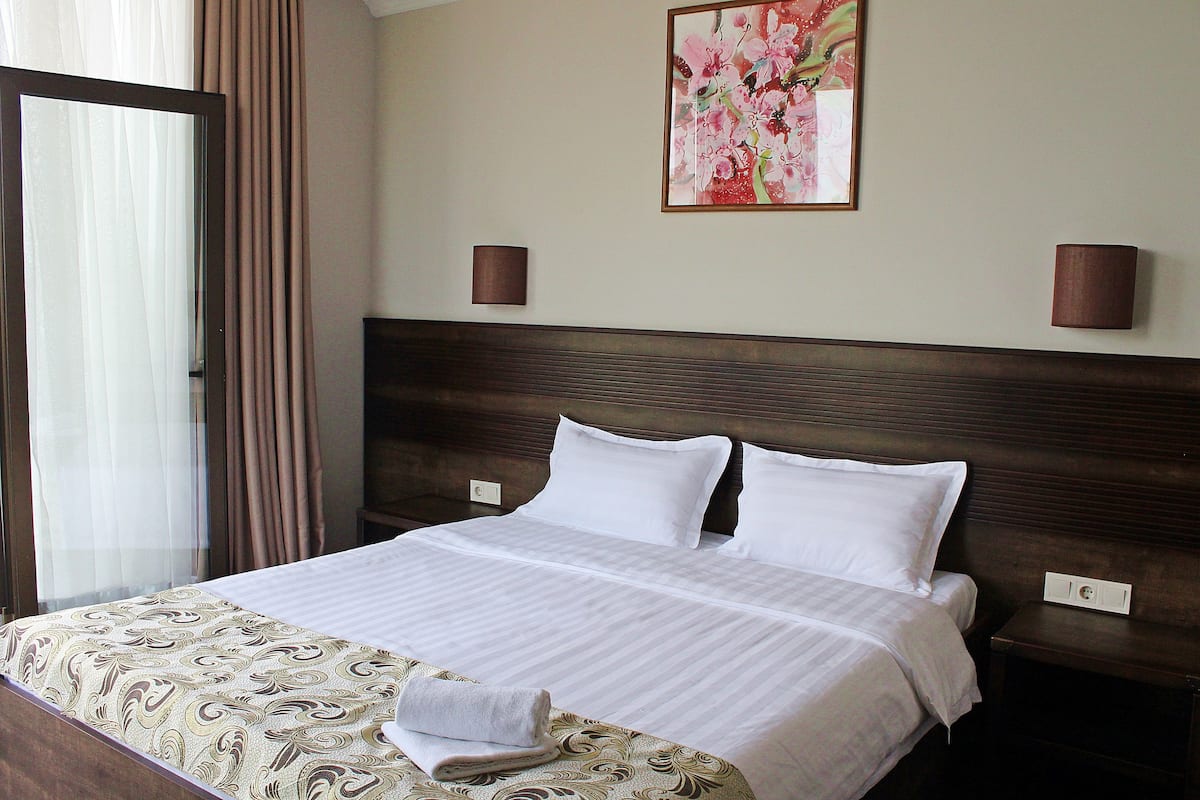 Habitación de hotel con una cama grande, dos mesitas de noche, un buró, una ventana con cortinas y un cuadro floral en la pared.