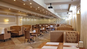 A restaurant with multiple dining tables and chairs, a projector on the ceiling, and decorative wall sconces.
