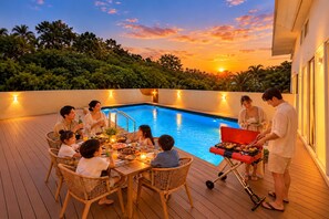 A group of people dining by a pool with a sunset in the background.